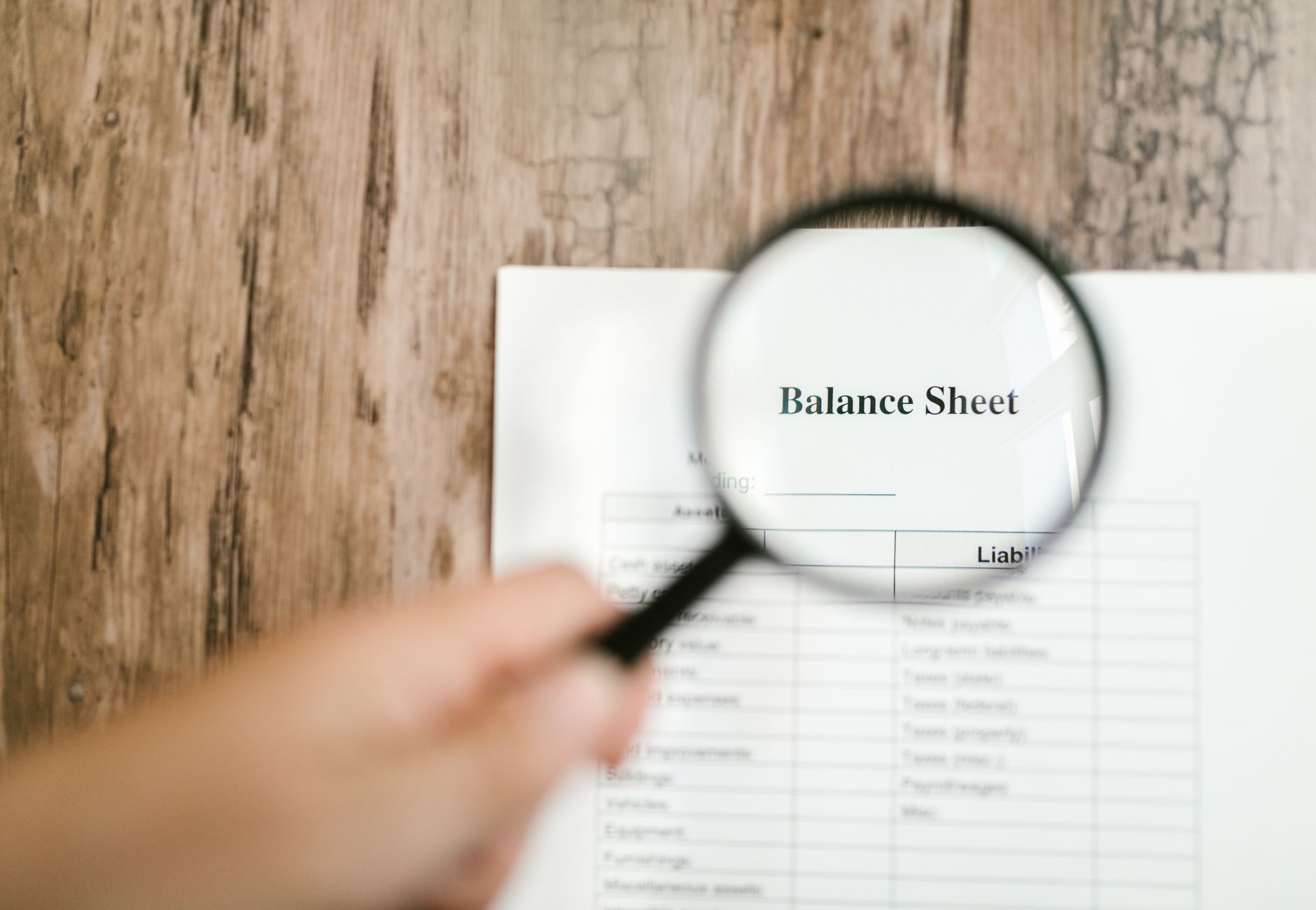 Close-up of a balance sheet document on wooden surface with a magnifying glass held by a hand.